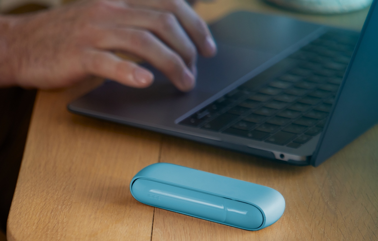 Hands typing on laptop and Iphone next to turquoise IQOS ORIGINALS DUO pocket charger.